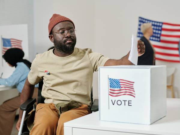 African American young man with disability sitting in wheelchair and putting ballot paper into box with USA flag standing on desk