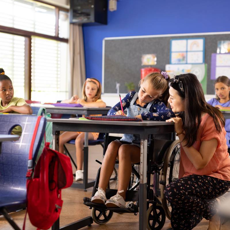 Diverse female teacher and schoolgirl in wheelchair in elementary school class