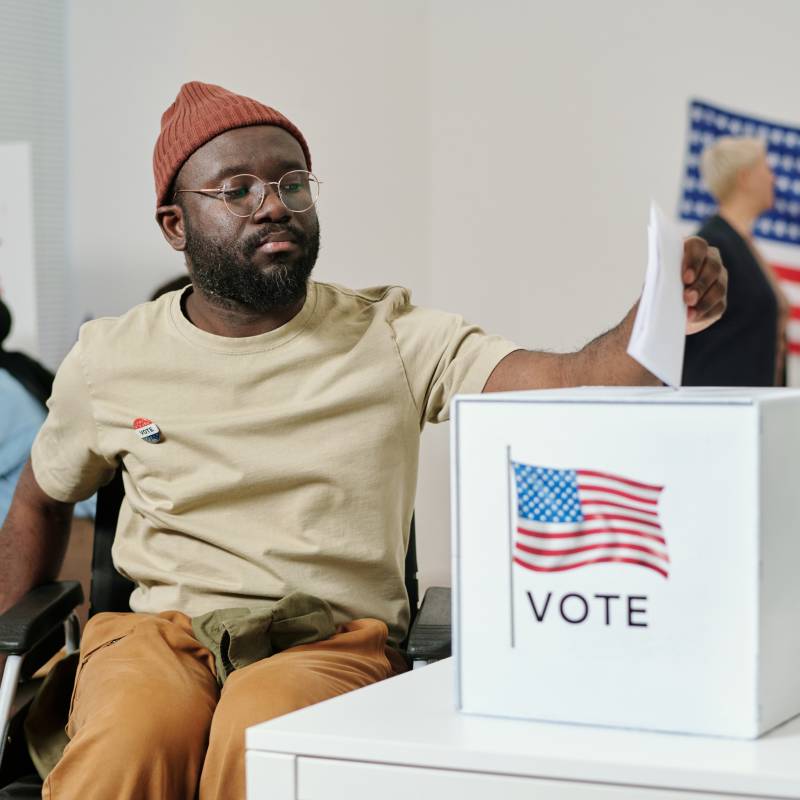 African American young man with disability sitting in wheelchair and putting ballot paper into box with USA flag standing on desk